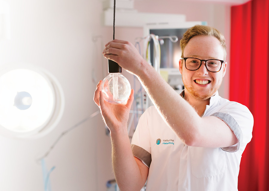 Een zorgprofessional van het Catharina Ziekenhuis toont lachend een grote gloeilamp in een lichte kamer met medische apparatuur.