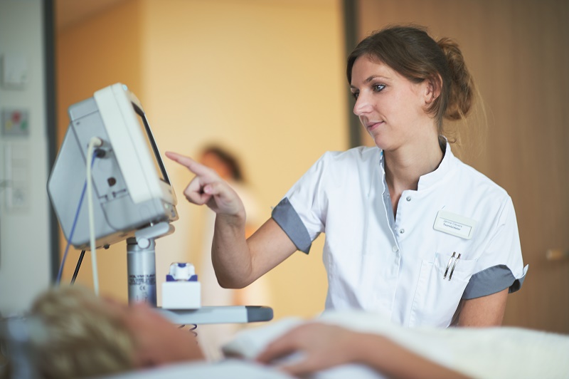 Zorgverlener in wit uniform bedient Step Forward Unit monitor naast patiënt in bed en drukt op knop ter ondersteuning van ontslag.