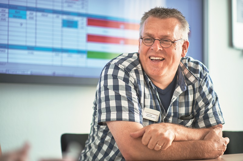 Man met bril en geruite blouse zit glimlachend aan tafel, draagt naamkaartje en stethoscoop. Op achtergrond kleurrig spreadsheet.