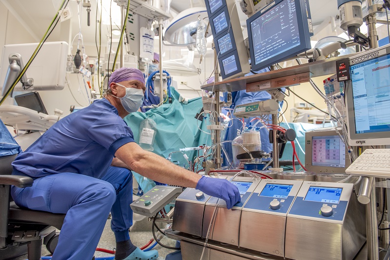 Een klinisch perfusionist in blauwe kleding bedient apparaten bij een bed, naast een patiënt onder een steriel laken.