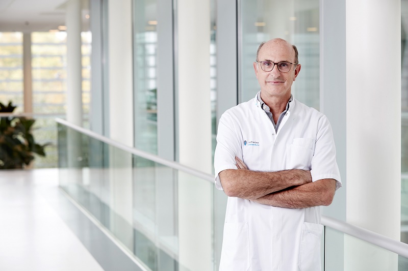 Een man met bril en witte jas staat met gekruiste armen in een lichte bende en steunt patiënten tijdens Darmkankermaand.