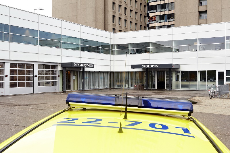 Een gele ambulance met blauwe lichtbalk staat voor een modern gebouw met ramen en borden DIENSTAPOTHEEK en SPOEDPOST.