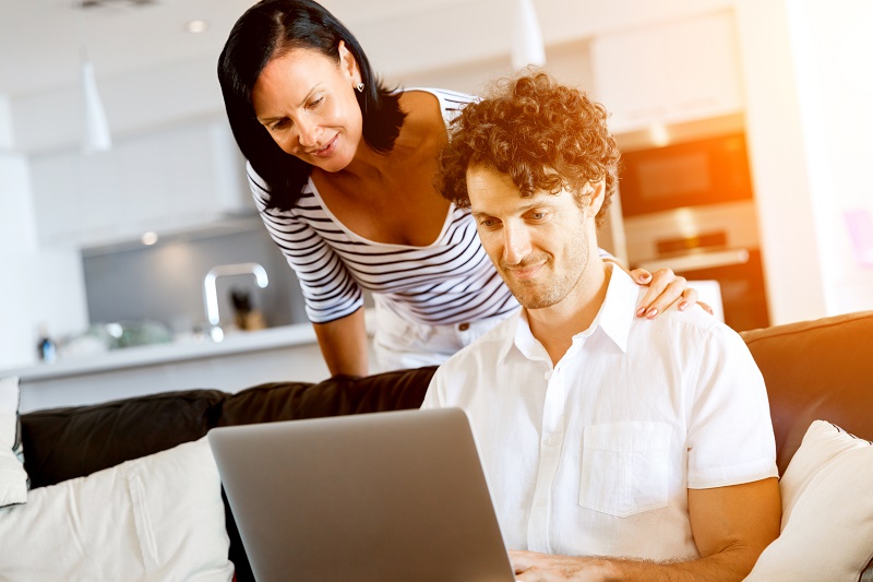 Man op bank gebruikt laptop, vrouw staat erachter met hand op zijn schouder; beiden kijken gefocust naar het scherm.
