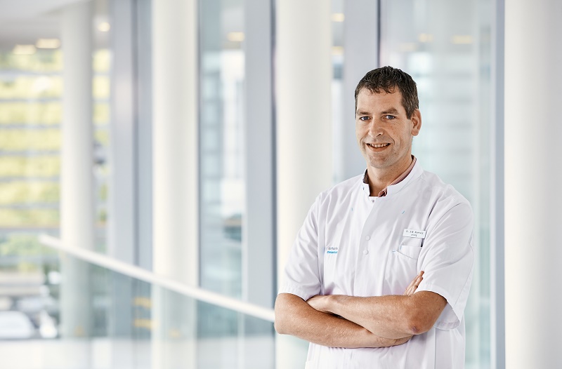 Man in witte doktersjas staat binnen met gekruiste armen en glimlach, in modern gebouw met grote ramen en witte pilaren.