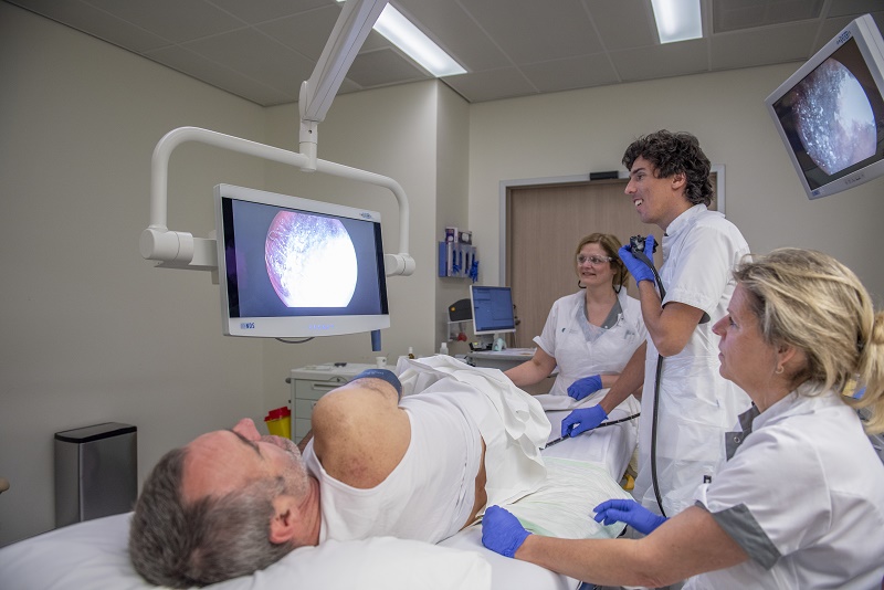 Patiënt op ziekenhuisbed, drie zorgverleners met witte jas en blauwe handschoenen kijken naar monitor bij colonoscopie.