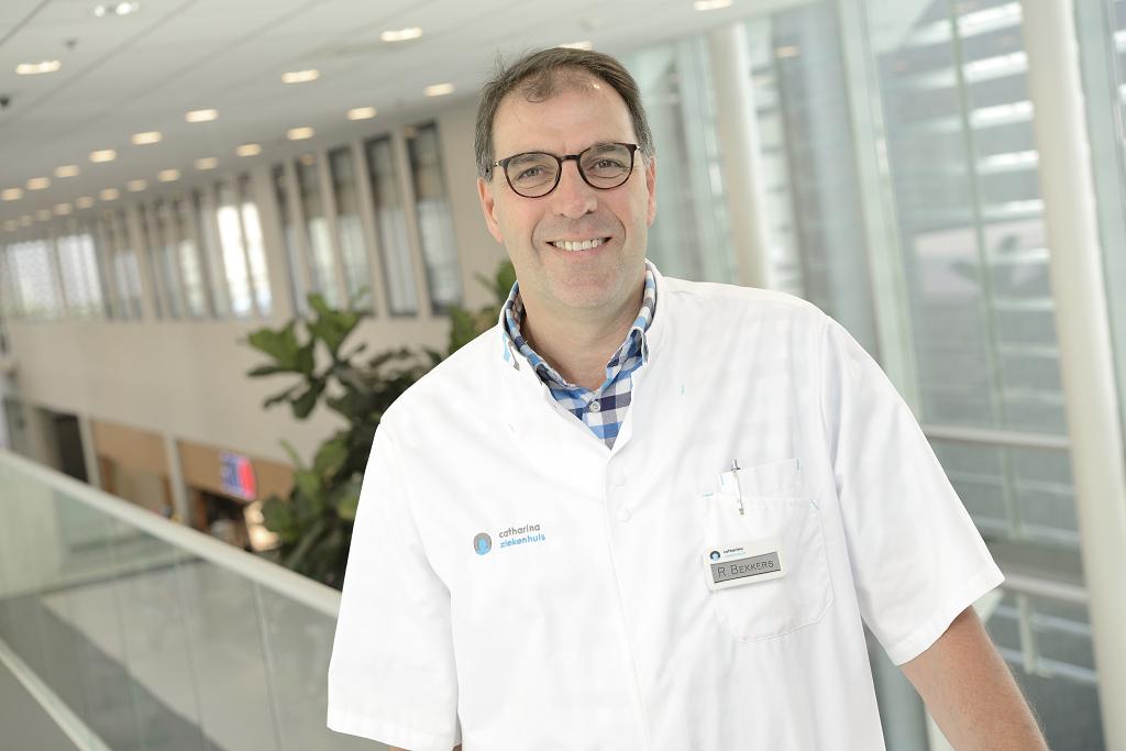 Een man met bril en witte labjas staat lachend binnen in een modern gebouw. Op de achtergrond zijn ramen, lampen en een plant.