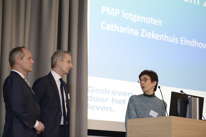 Drie mensen voorin de zaal; twee mannen in pak tegenover een vrouw bij een spreekgestoelte, scherm met tekst over PMP-lotgenoten.