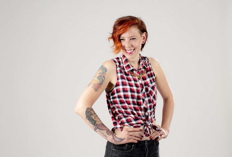 Persoon met kort rood haar, mouwloos ruitjeshemd, zwarte broek en tatoeages poseert glimlachend tegen lichte achtergrond.