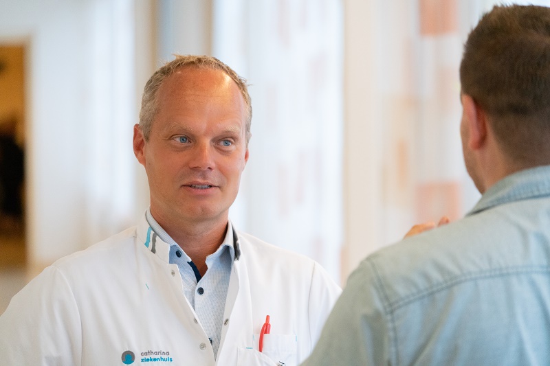 Een chirurg in witte jas spreekt met iemand in de bende; de tweede persoon is van achteren te zien.