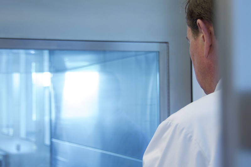 Persoon in witte labjas met rug naar camera kijkt in grote glazen ruimte met blauwe tint in het laboratorium van Catharina Ziekenhuis.