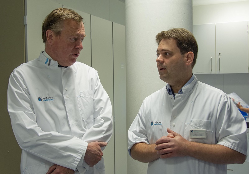 Twee mannen in witte jassen praten binnen in het Catharina Ziekenhuis, met kasten en witte muren op de achtergrond.