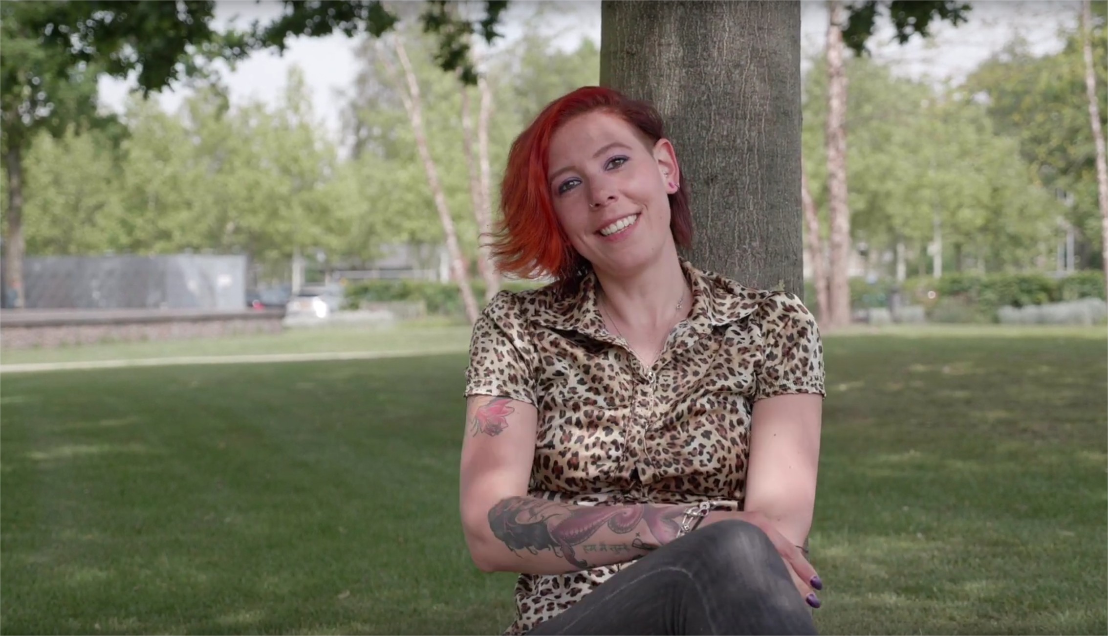 Persoon met kort rood haar zit in het gras tegen een boom, draagt luipaardprint en heeft tattoos op beide armen.
