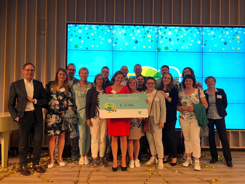 Groep mensen bij scherm met blauwe achtergrond en sterren. Twee vrouwen tonen cheque van €10.000. Goud confetti op vloer.