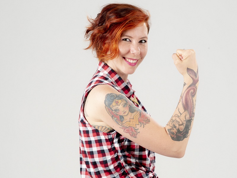 Persoon met kort rood haar, mouwloos ruitjeshemd, toont getatoeëerde arm en lacht voor blogfoto, lichte achtergrond.