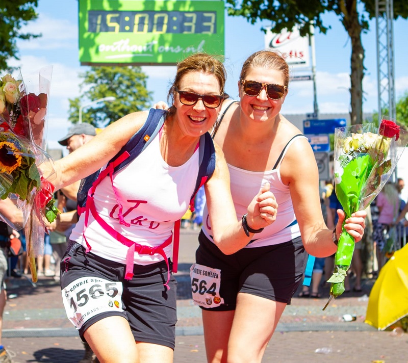 Twee vrouwen in sportkleding met bloemen poseren bij de loop voor kanker. Ze dragen startnummers en een timer is zichtbaar.