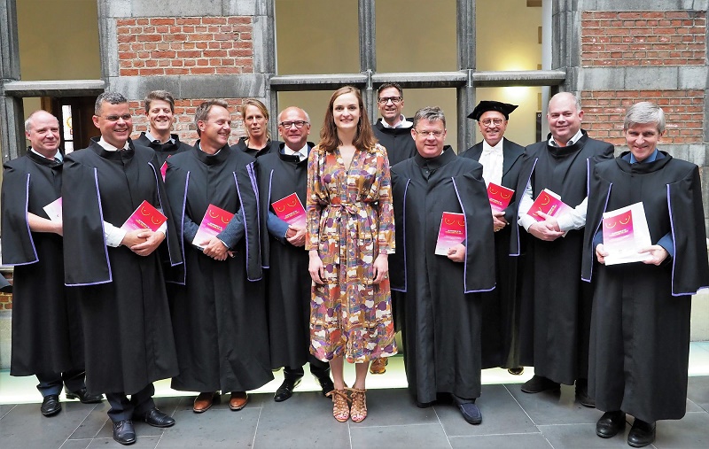 Twaalf volwassenen, één in jurk met patroon en elf in zwarte toga's, poseren binnen met rode boekjes na borstkankeroperatie.