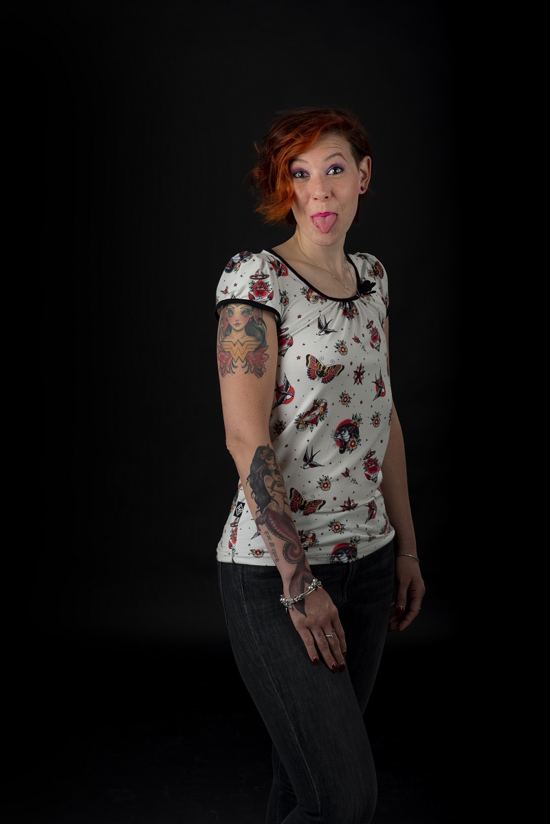 Persoon met kort rood haar, patroonshirt en donkere jeans, steekt tong uit, tattoos op rechterarm, zwarte achtergrond.