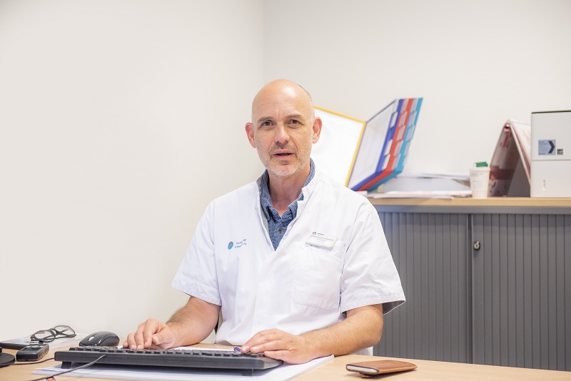 Gynaecoloog-oncoloog Ralph Hermans typt aan een bureau in een lichte kamer, met kantoorartikelen en koffiebeker achter hem.