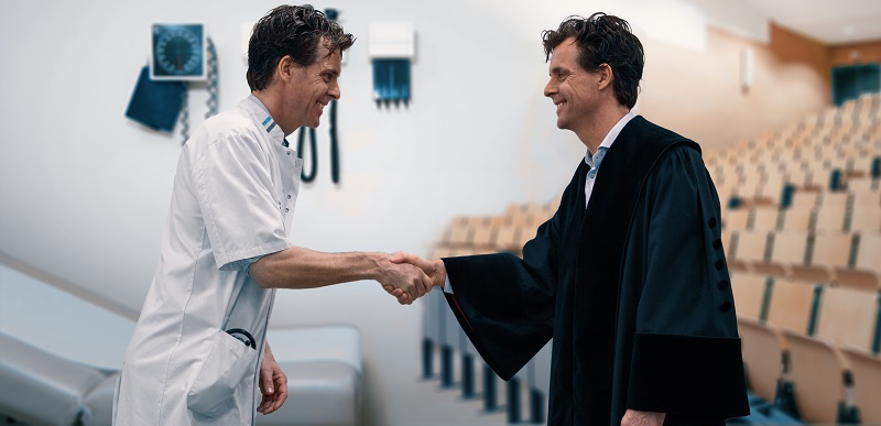 Een arts in wit uniform schudt handen met hoogleraar Lukas Dekker in zwarte toga, in een lege collegezaal.