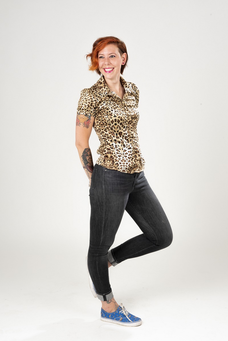 Naomi van moedertoekomst staat op één been met glimlach, handen op het tapijt. Ze heeft rood haar, panterblouse, jeans en blauwe sneakers.