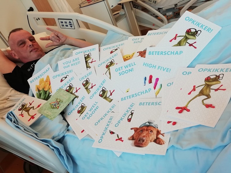 Erik ligt in een ziekenhuisbed en maakt een peace-teken, omringd door kleurrijke opkikkerkaarten. Zakdoekje op bed.