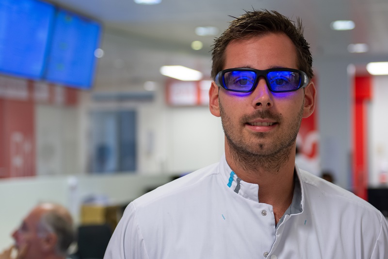 Man met blauwe veiligheidsbril en witte labjas in Catharina Ziekenhuis; op achtergrond computers en persoon, mogelijke nachtdienst.
