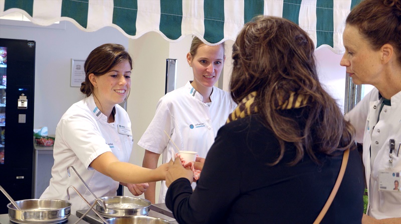 Twee diëtisten in witte uniformen schenken soep aan een persoon bij een balie, onder een gestreepte luifel in het Catharina Kanker Instituut.