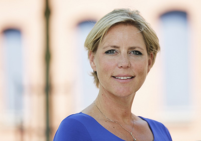 Vrouw met kort blond haar en blauw shirt staat buiten voor een gebouw met grote ramen. Ze kijkt in de camera en glimlacht.