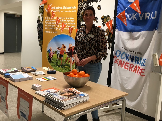 Vrouw achter informatietafel met folders, kaarten en sinaasappels; banners promoten rookvrij ziekenhuis en stoppen-met-roken tips.