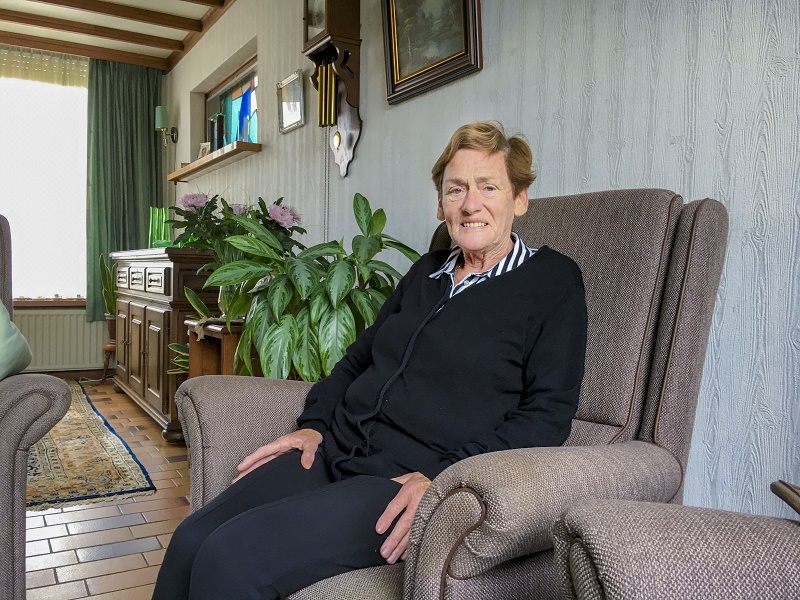 Oudere vrouw met kort haar en zwarte trui zit in een bruine stoel thuis, met planten en schilderijen op de achtergrond.