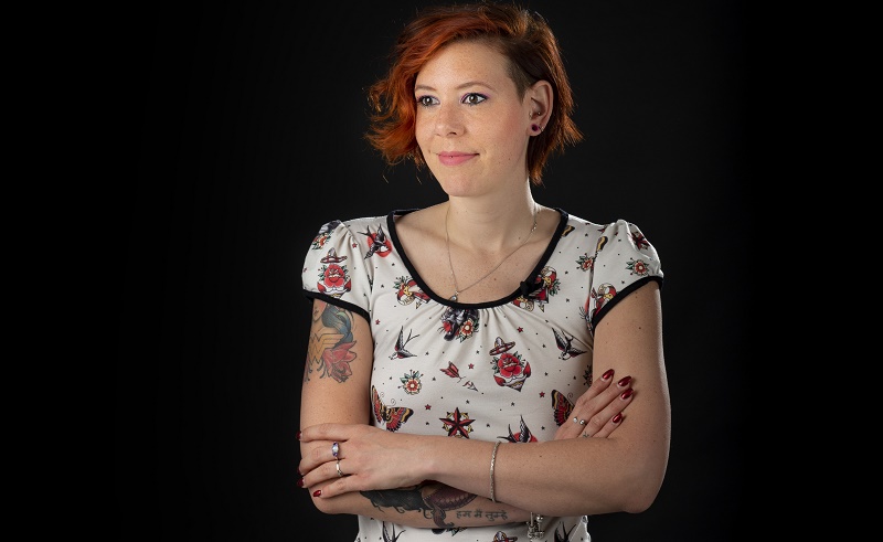 Persoon met kort rood haar en tattoos op beide armen, gekruiste armen, wit shirt met kleurrijke patronen voor zwarte achtergrond.