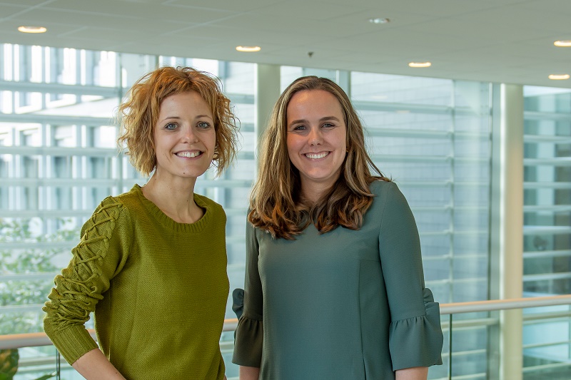 Twee vrouwen staan binnen voor grote ramen met uitzicht op een modern bankgebouw. Ze dragen groene toppen en glimlachen.