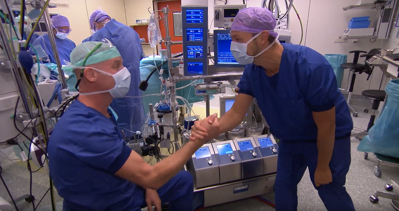Twee medici in OK-kleding geven elkaar een hand tijdens een kloppend hart procedure; collega's werken op de achtergrond.