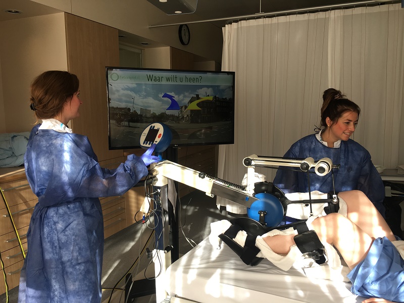 Twee vrouwen bedienen een revalidatierobot aan het been van een patiënt naast een ziekenhuisbed; monitor toont Nederlands.