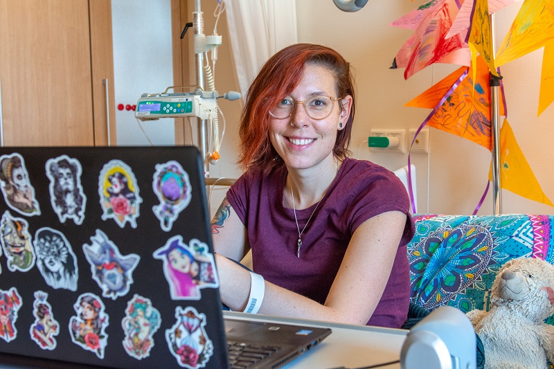 Persoon met kort rood haar en bril werkt op een laptop in een lichte ziekenhuiskamer. Apparatuur en speelgoed op de achtergrond.