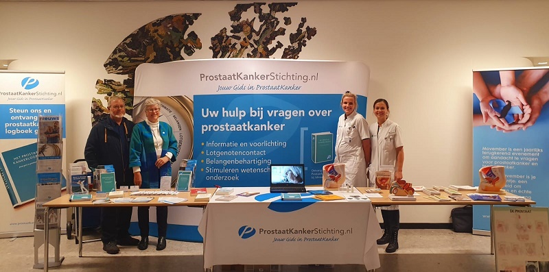 Vier mensen bij een prostaatkankerstand met blauwe banners, folders, modellen en scherm. Promotie van ProstaatKankerStichting.nl.