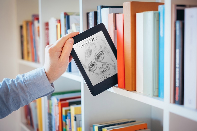 Hand houdt een e-reader met een getekend lachend gezicht, voor een boekenkast. Op het scherm staat "Laatje me dan".