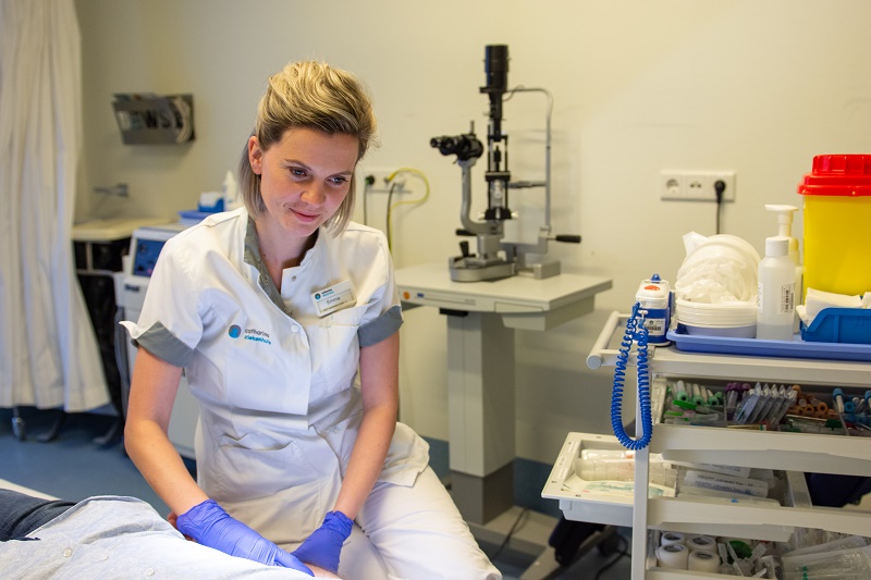 Een zorgmedewerker in wit uniform en blauwe handschoenen zit naast een patiënt in een onderzoeksruimte met medische apparatuur.