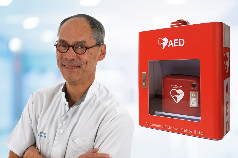 Man in witte zorgkleding en bril bij rode AED aan muur, met wazige achtergrond van een medische faciliteit.