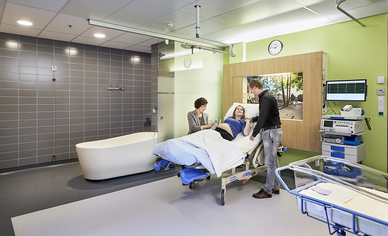 Vrouw ligt in ziekenhuisbed bij Vrouw Kind Centrum, samen met ouders en kind, omringd door medische apparatuur en een wieg.