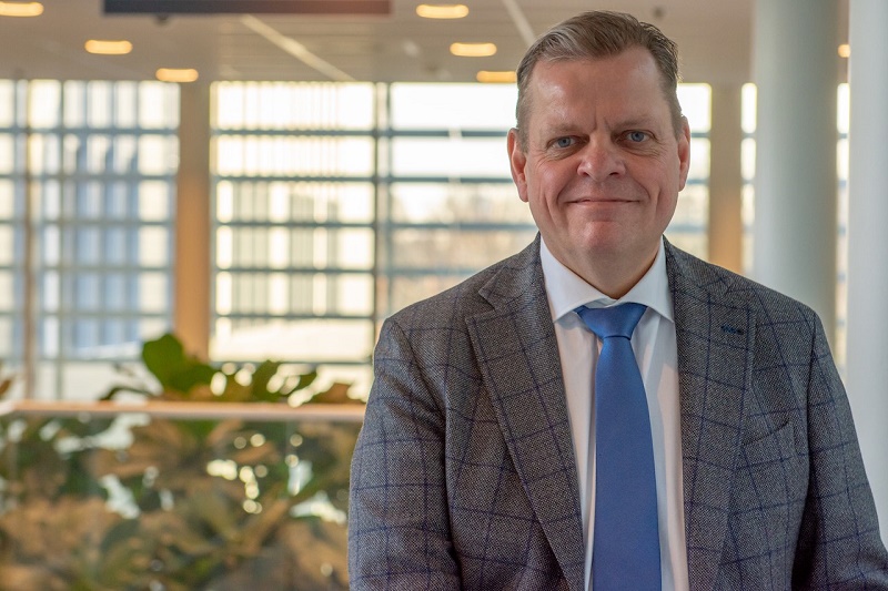 Een man van middelbare leeftijd in geruit colbert, blauwe stropdas en wit overhemd staat binnen bij grote ramen en planten.