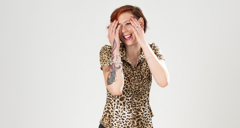 Persoon met kort rood haar en tatoeages op linkerarm glimlacht, bedekt deels gezicht, draagt panterprint shirt, effen achtergrond.