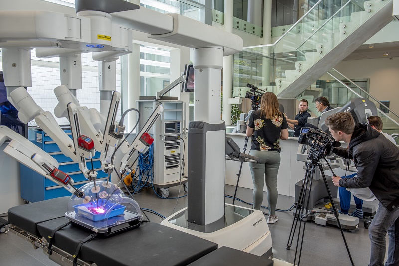 Groep mensen filmt demonstratie van een medisch robotsysteem met robotarmen boven een patiëntsimulator op operatietafel.