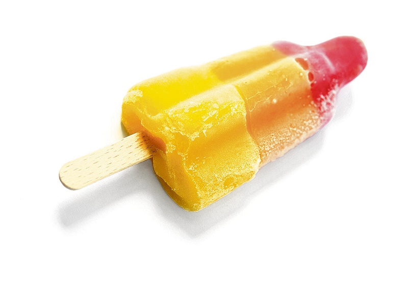 Een geel-rood waterijsje ligt op zijn zij op een witte ondergrond, met het houten stokje deels zichtbaar. Het ijsje rook.