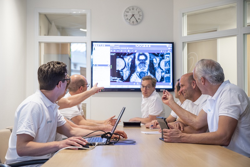 Vijf mannen in witte hemden bespreken prostaatkankerbeelden op een groot scherm aan een vergadertafel in een centrum.