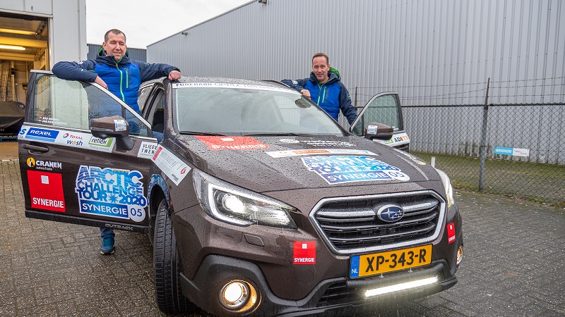 Twee mannen bij een donkere Subaru met Arctic Challenge Tour-stickers en NL-kenteken, geparkeerd bij een industrieel pand.