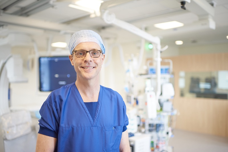 Persoon in blauwe scrubs, operatiemuts en bril staat in modern Eindhovens ziekenhuis met medische apparatuur op de achtergrond.