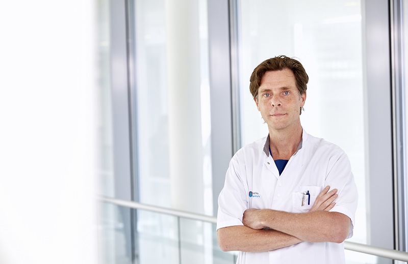 Een persoon in een witte artsenjas staat met gekruiste armen in een lichte gang met glazen wanden; mogelijk Lukas Dekker.