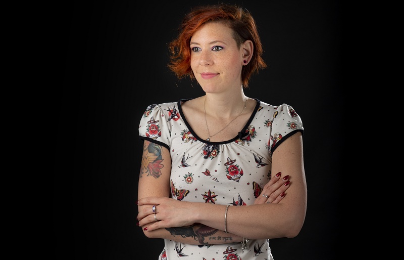 Persoon met kort rood haar en tatoeages staat met gekruiste armen in licht shirt met ingrediëntenpatroon voor zwarte achtergrond.