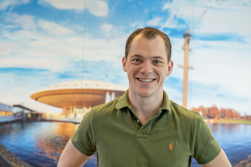 Man in groen poloshirt glimlacht voor futuristisch rond gebouw aan het water onder blauwe lucht met wolken.